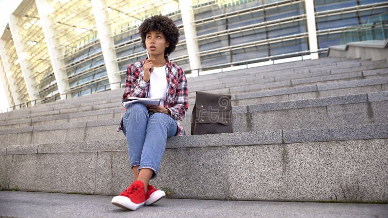 Estudiante cabelludo rizado lindo que hace la preparaci?n, sent?ndose en las escaleras cerca de arena imagenes de archivo Estudiante cabelludo rizado lindo que hace la preparaci?n, sent?ndose en las escaleras cerca de arena imagenes de archivo