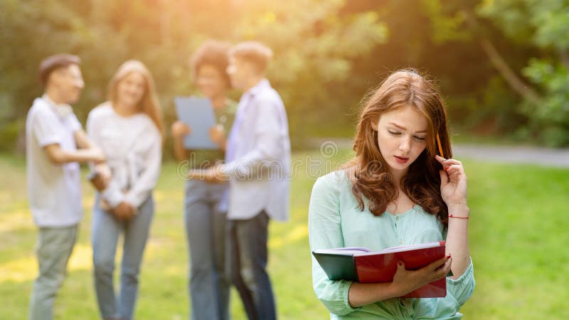 Estudiante pelirroja centrada en la preparación para una conferencia al aire libre mientras sus compañeros charlan fotos de archivo libres de regalías
