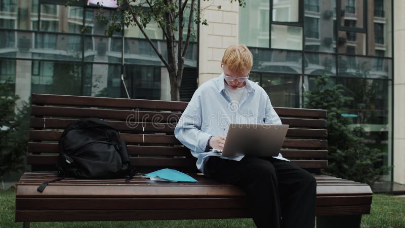 Estudiante se dedicó a escribir con enfoque en un portátil mientras estaba sentado en un banco al aire libre almacen de video