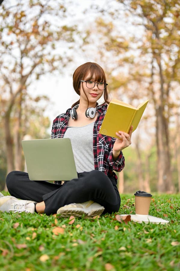 Portrait,Focused and attractive young Asian female college student sits on the grass in the park,reads a book,and uses her laptop to manage her school work. uni-lifestyle concept. Portrait,Focused and attractive young Asian female college student sits on the grass in the park,reads a book,and uses her laptop to manage her school work. uni-lifestyle concept