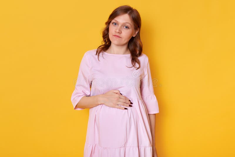 Estudio tirado de la mujer embarazada bonita joven que lleva el vestido rosado del polvo y que lleva a cabo la mano en el vientre fotografía de archivo Estudio tirado de la mujer embarazada bonita joven que lleva el vestido rosado del polvo y que lleva a cabo la mano en el vientre fotografía de archivo