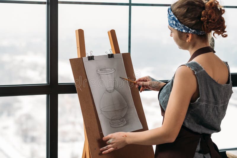Art class. Side view of young lady drawing sketch of vase in studio. French window background. Art class. Side view of young lady drawing sketch of vase in studio. French window background.