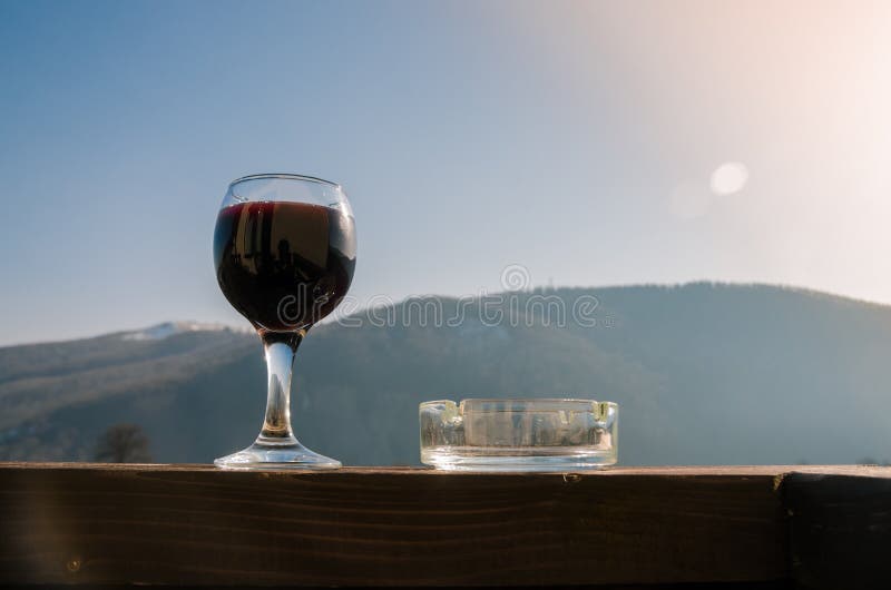 Ett exponeringsglas av rött vin och ett askfat på bakgrund av berg royaltyfri bild Ett exponeringsglas av rött vin och ett askfat på bakgrund av berg royaltyfri bild