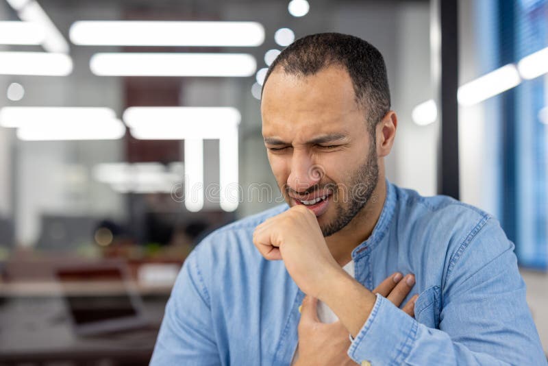 Close-up photo of a young sick hispanic man sitting in the office and coughing covering his mouth with his hand and holding his chest. Close-up photo of a young sick hispanic man sitting in the office and coughing covering his mouth with his hand and holding his chest.