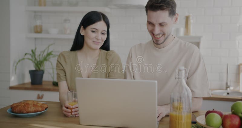 Ett lyckligt ungt par som ringer videosamtal online med en bärbar dator som pratar och viker hand i köket hemma