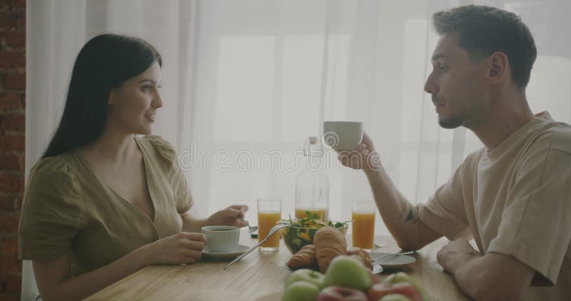 Ett lyckligt ungt par som äter frukost och dricker kaffe och som sitter på bordet hemma i köket