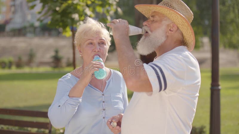 Ett medelålders kaukasiskt par som dricker smakligt vatten i sommarparken och som chattar Positiv mogen man i halmhatt