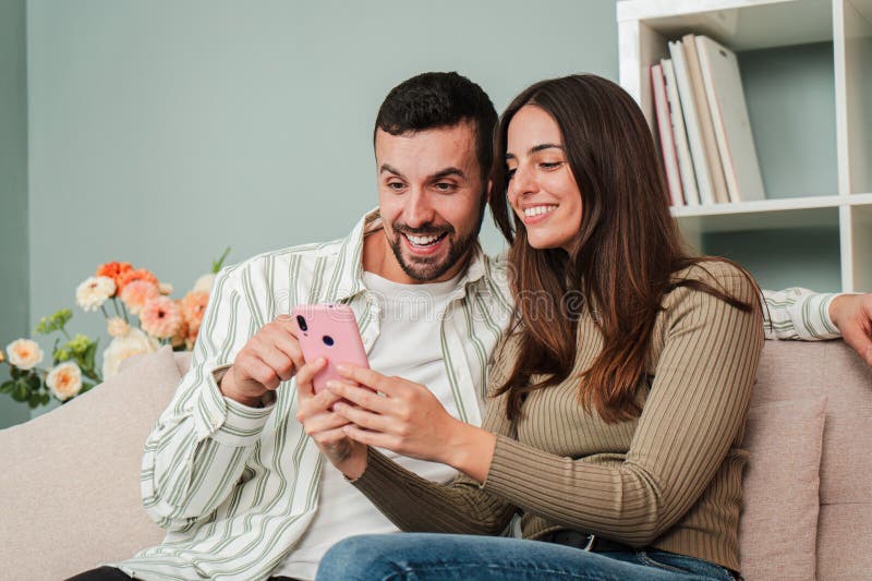 Caucasian couple having fun using a mobile phone to browse on internet at living room sofa. Two friends betting online with a cellphone app. Boyfriend and girlfriend searching or booking an apartment. High quality photo. Caucasian couple having fun using a mobile phone to browse on internet at living room sofa. Two friends betting online with a cellphone app. Boyfriend and girlfriend searching or booking an apartment. High quality photo