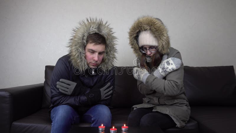 A couple of young people are sitting in jackets at home and warming themselves from candles. A man and a woman have not paid their heating bills and are freezing at home. 4k. A couple of young people are sitting in jackets at home and warming themselves from candles. A man and a woman have not paid their heating bills and are freezing at home. 4k