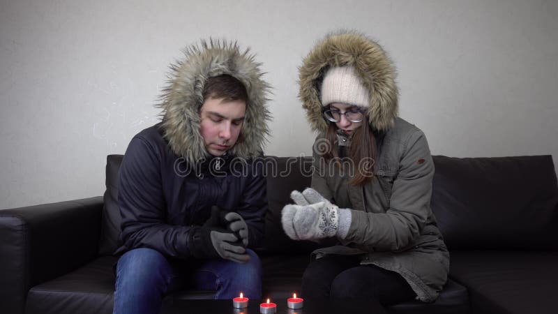 A couple of young people are sitting in jackets at home and warming themselves from candles. A man and a woman have not paid their heating bills and are freezing at home. 4k. A couple of young people are sitting in jackets at home and warming themselves from candles. A man and a woman have not paid their heating bills and are freezing at home. 4k