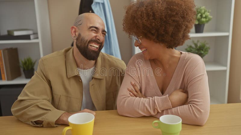 Ett skrattretande interrasistiskt par som har en tillfällig inomhusmiljö med kaffekoppor på bordet royaltyfri bild