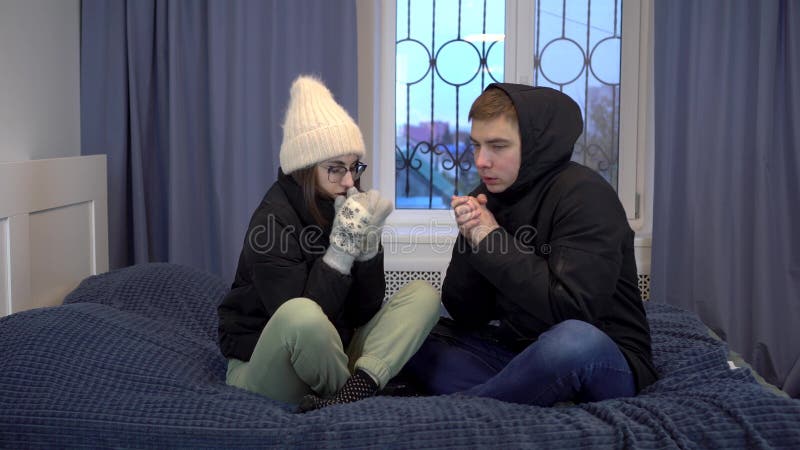 A young couple sit in jackets at home hugging to keep warm. Turning off the heating due to expensive builds. They sit in the room and get cold. 4k. A young couple sit in jackets at home hugging to keep warm. Turning off the heating due to expensive builds. They sit in the room and get cold. 4k