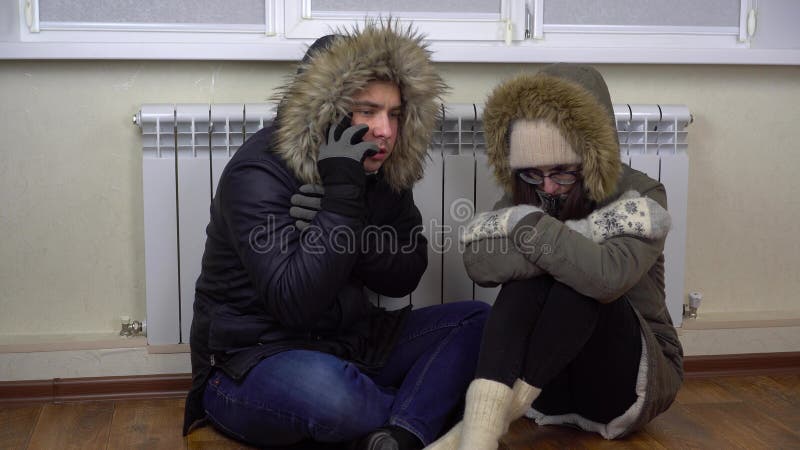 A young couple is sitting on the floor by the radiator in winter jackets. A man calls on the phone in a company. to turn on the heating. 4k. A young couple is sitting on the floor by the radiator in winter jackets. A man calls on the phone in a company. to turn on the heating. 4k