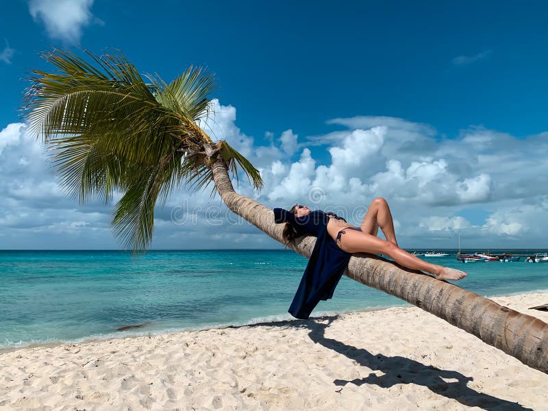 European beautiful happy dark haired girl with sunglasses in bikini and blue pareo is lying on coconut palm on the white sand
