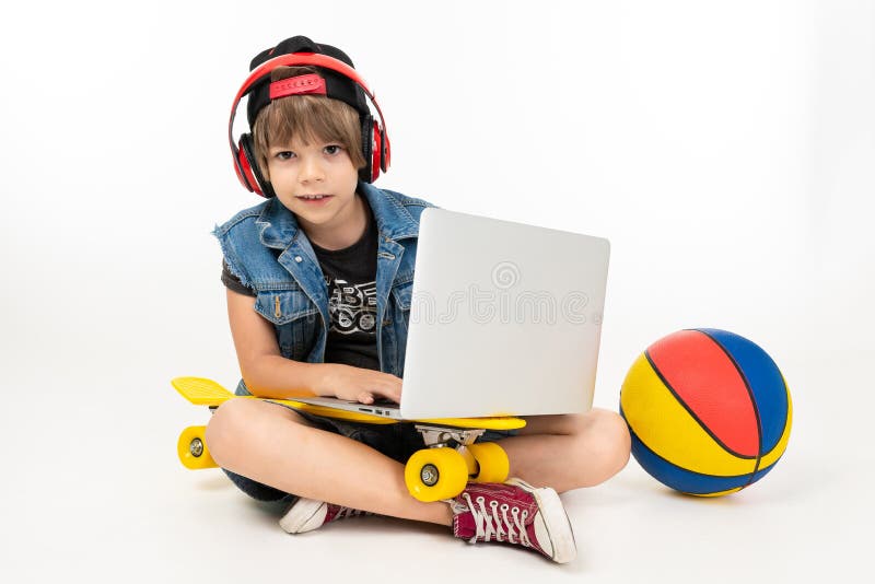 European boy sitting on the floor with laptop, skateboard and basketball on a white background.