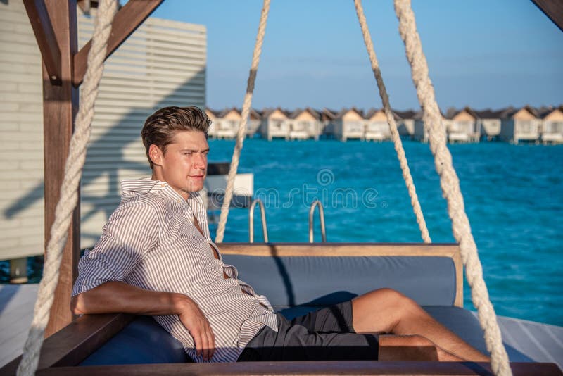 European handsome man wearing shirt and shorts relaxing on the sofa at tropical beach at island luxury resort in Maldives