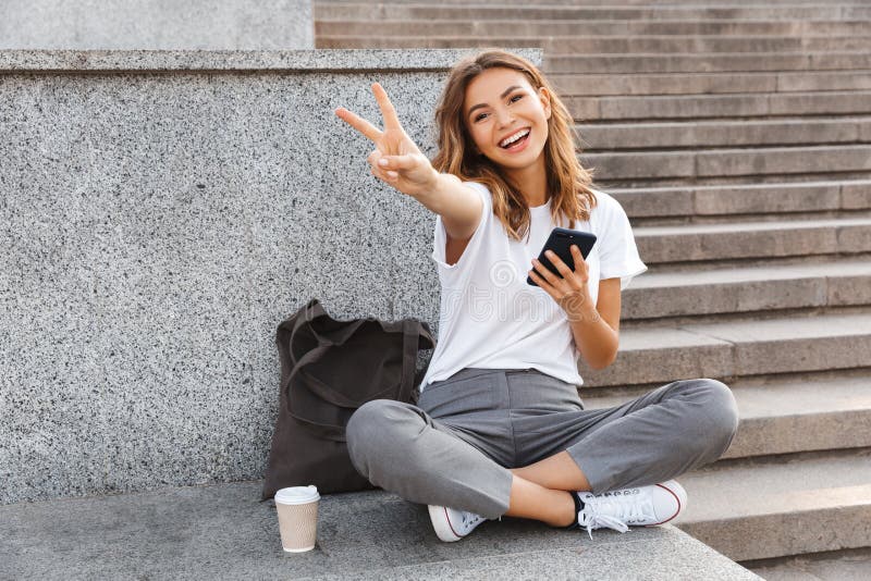 European smiling woman sitting on street stairs with legs crossed on summer day, and showing peace sign with mobile phone in hand royalty free stock image European smiling woman sitting on street stairs with legs crossed on summer day, and showing peace sign with mobile phone in hand royalty free stock image
