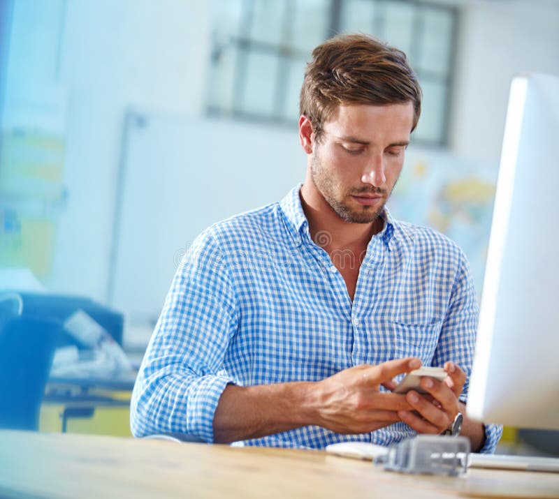 Everything is done via text these days. a designer using a cellphone while working on a computer in an office.