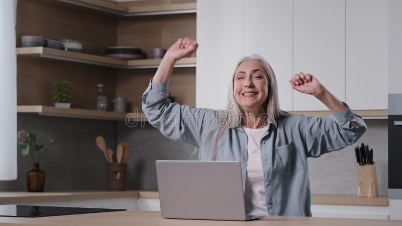 Excitada mulher idosa ganhadora de seniores impressionada mulher de negócios sentada na cozinha celebra vitória vence vendas onlin