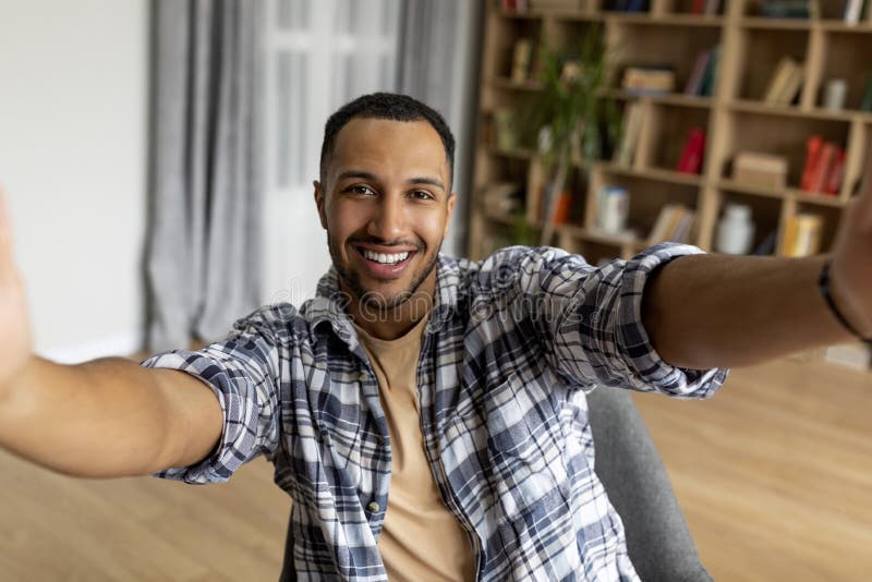 Excited arab guy taking selfie and smiling at mobile phone camera, sitting in armchair at home, free space stock photo
