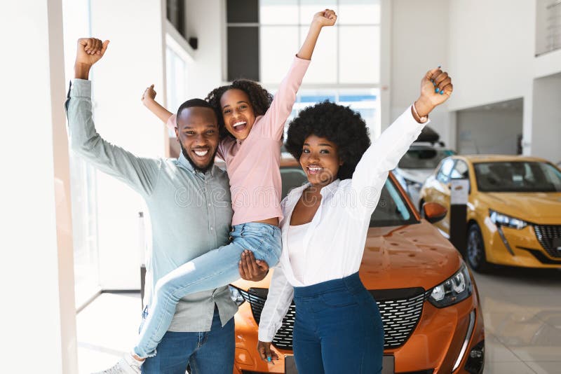 Excited black family of three lifting hands up, celebrating new car purchase in auto dealership. Young parents with daughter gesturing YES after buying vehicle at automobile showroom