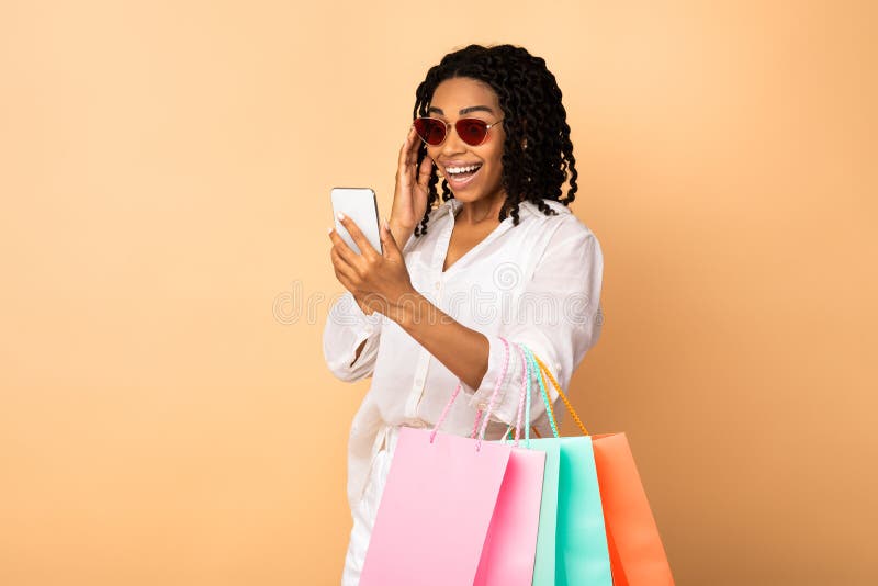 Excited Black Lady Using Phone Holding Shopper Bags, Beige Background