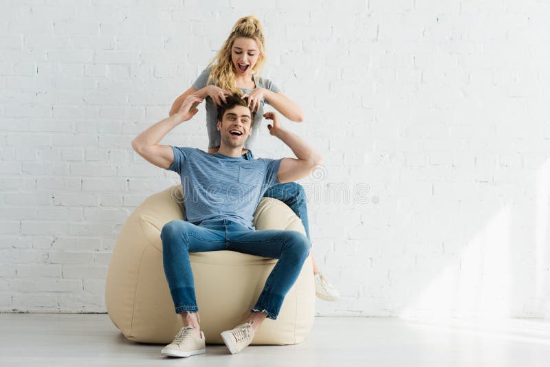 Excited blonde girl touching hair of happy men sitting on beige bean bag chair at home. Excited blonde girl touching hair of happy men sitting on beige bean bag chair at home