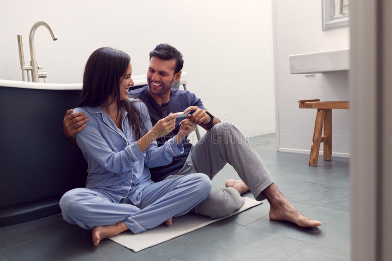 Excited Couple Sitting On Floor In Bathroom At Home With Positive Home Pregnancy Test Stock