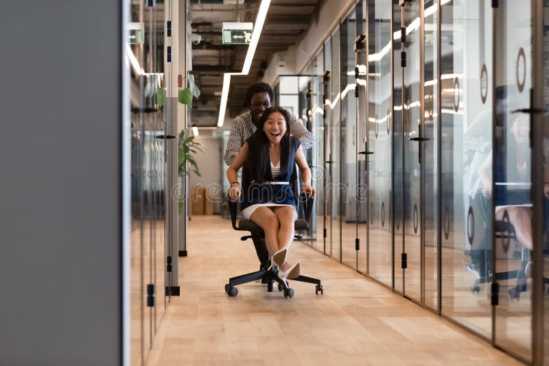 Excited diverse colleagues laughing riding on chair in office hallway