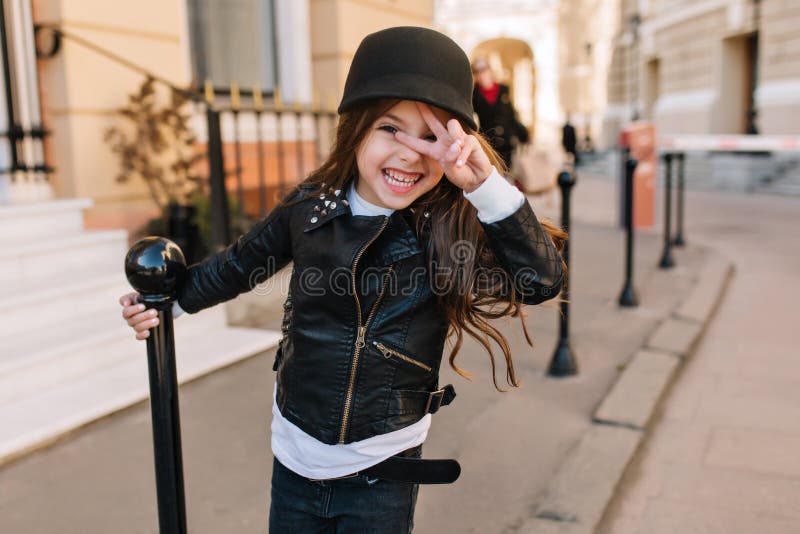 Excited little girl wearing leather jacket and belt holding iron pillar and posing with peace sign on city background royalty free stock photography Excited little girl wearing leather jacket and belt holding iron pillar and posing with peace sign on city background royalty free stock photography