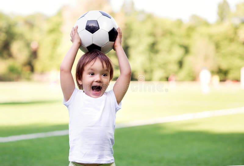 Little toddler boy playing football on soccer field outdoors: the kid is holding ball above head and shouting ready to throw it royalty free stock photo Little toddler boy playing football on soccer field outdoors: the kid is holding ball above head and shouting ready to throw it royalty free stock photo
