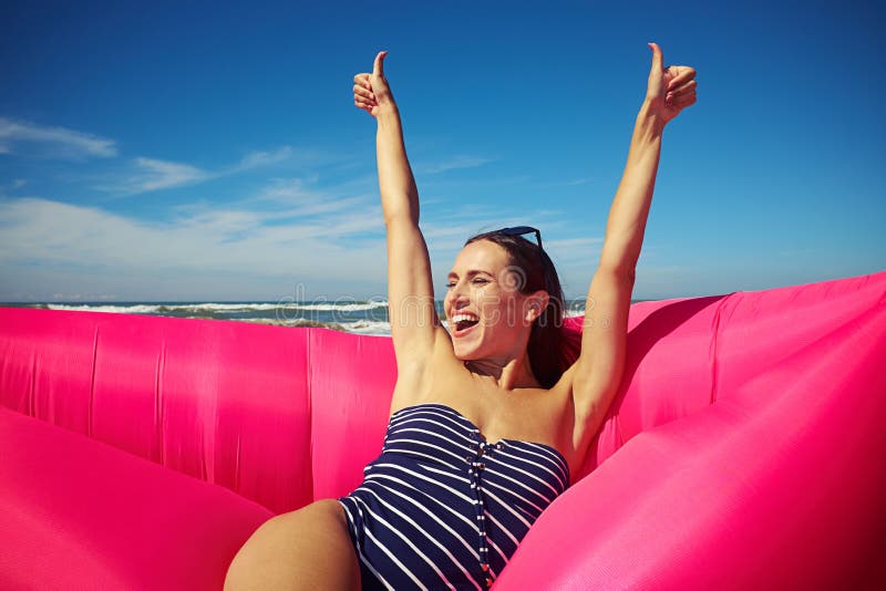 Mid shot of excited woman sitting on the rubber raft on the beach and holding her thumbs-up.and smiling. She’s wearing a striped swimming suit and dark sunglasses, her hair is put in a braid. A sunny day, blue sky and raging sea. Mid shot of excited woman sitting on the rubber raft on the beach and holding her thumbs-up.and smiling. She’s wearing a striped swimming suit and dark sunglasses, her hair is put in a braid. A sunny day, blue sky and raging sea