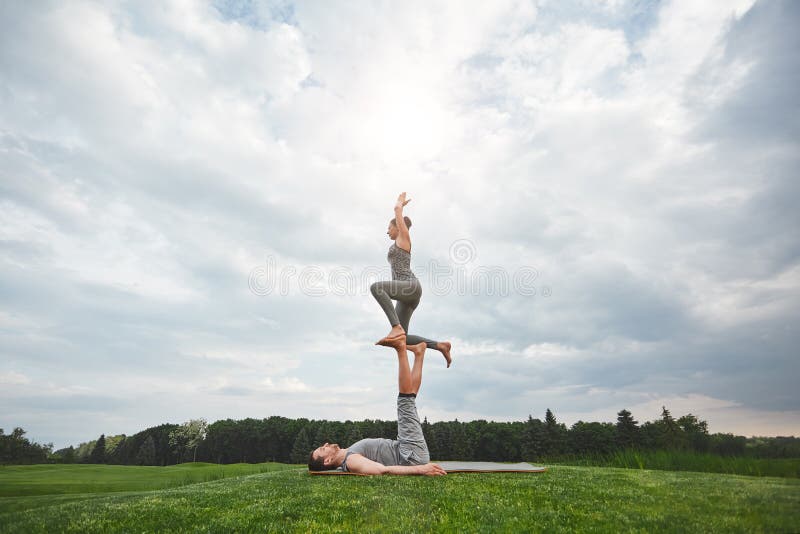 Exercices d'équilibre Couples sains et jeunes faisant le yoga acrobatique en nature Homme fort se trouvant sur un tapis et un équ photographie stock libre de droits Exercices d'équilibre Couples sains et jeunes faisant le yoga acrobatique en nature Homme fort se trouvant sur un tapis et un équ photographie stock libre de droits