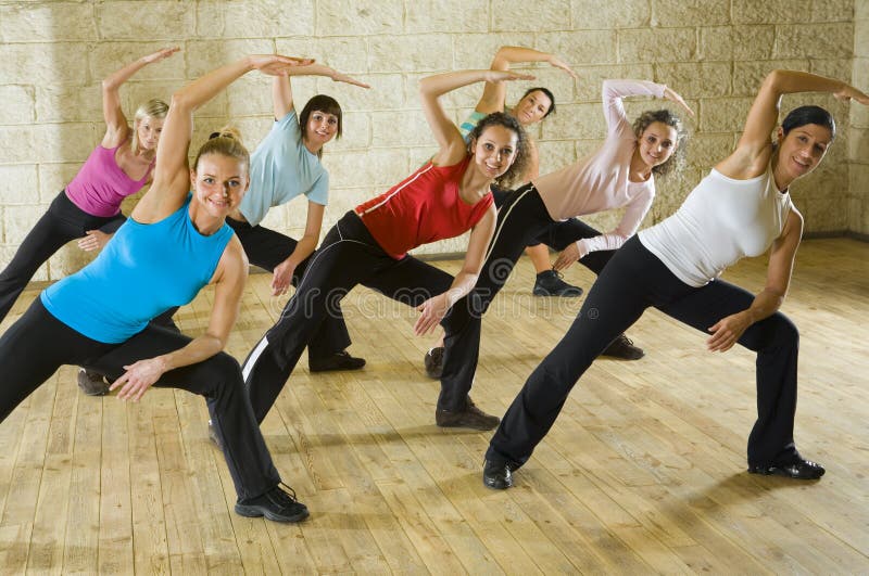 A group of women working out in the fitness club making stretching exercise. They're smiling and looking at camera. Front view. A group of women working out in the fitness club making stretching exercise. They're smiling and looking at camera. Front view.
