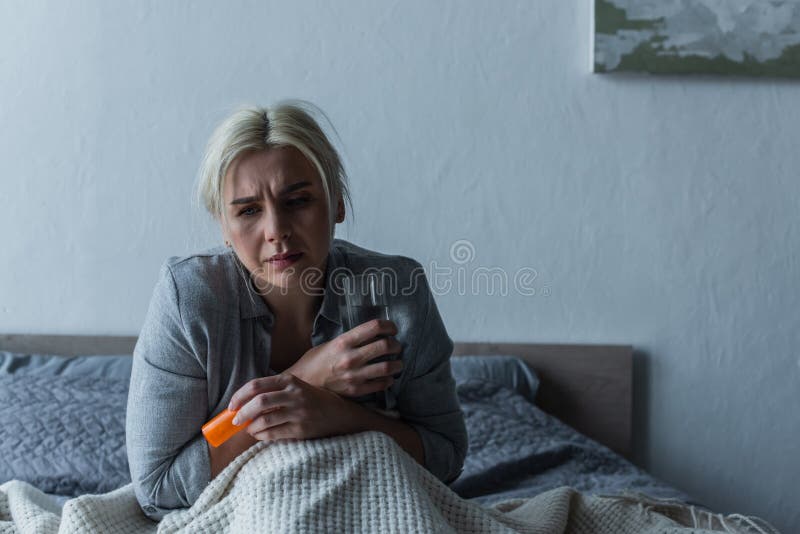 exhausted woman with climax sitting in bed while holding glass of water and painkillers,stock image