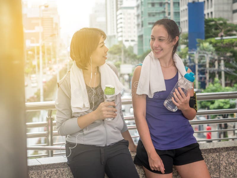 Exhausted after workout. Attractive young woman in sportswear drinking water while standing outdoor. They smile to each other stock image Exhausted after workout. Attractive young woman in sportswear drinking water while standing outdoor. They smile to each other stock image