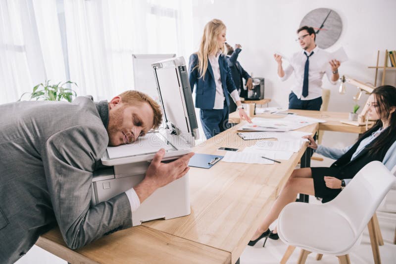 Exhausted zombie like businessman sleeping on copier while his colleagues having conversation