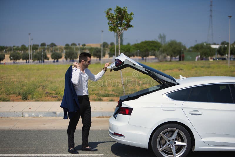Exitoso joven y atractivo hombre hispano con camisa blanca y chaqueta de hombro y gafas de sol abriendo el baúl de su blanco imágenes de archivo libres de regalías