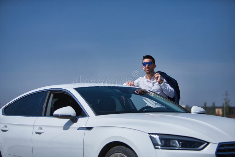 Exitoso joven y atractivo hombre hispano con camisa blanca y chaqueta sobre su hombro y gafas de sol apoyándose en el techo de fotos de archivo