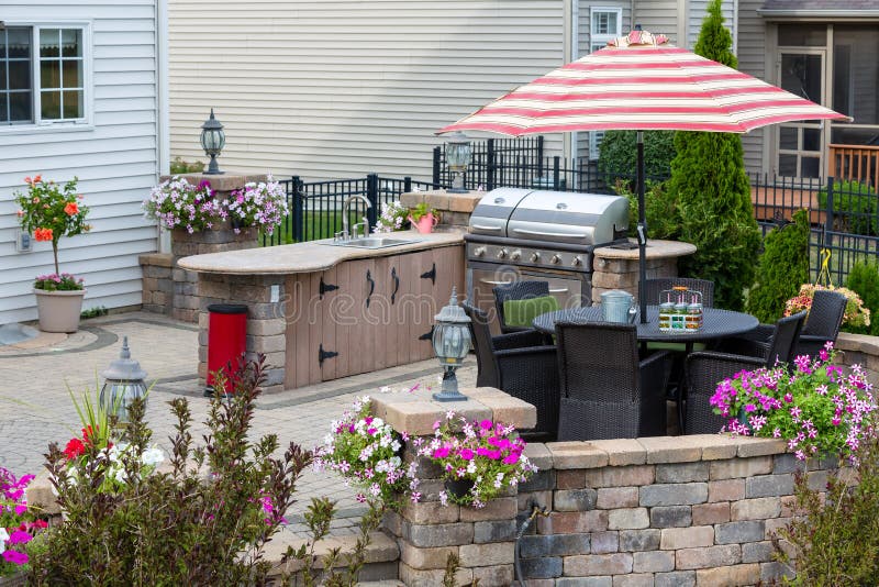 Upscale outdoor patio with kitchen area and comfortable wicker furniture in the shade of a striped umbrella surrounded by summer flowers. Upscale outdoor patio with kitchen area and comfortable wicker furniture in the shade of a striped umbrella surrounded by summer flowers