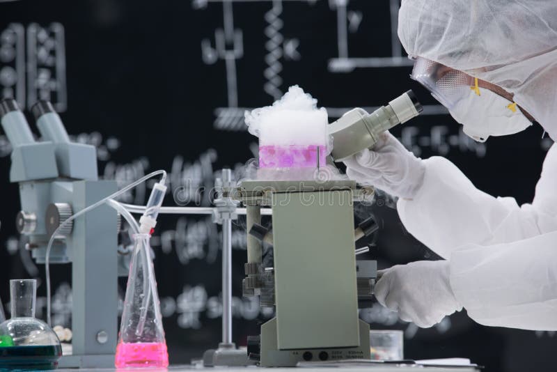 Close-up of scientist in a chemistry lab analyzing under microscope around colorful liquids and lab tools with a blackboard on the background. Close-up of scientist in a chemistry lab analyzing under microscope around colorful liquids and lab tools with a blackboard on the background