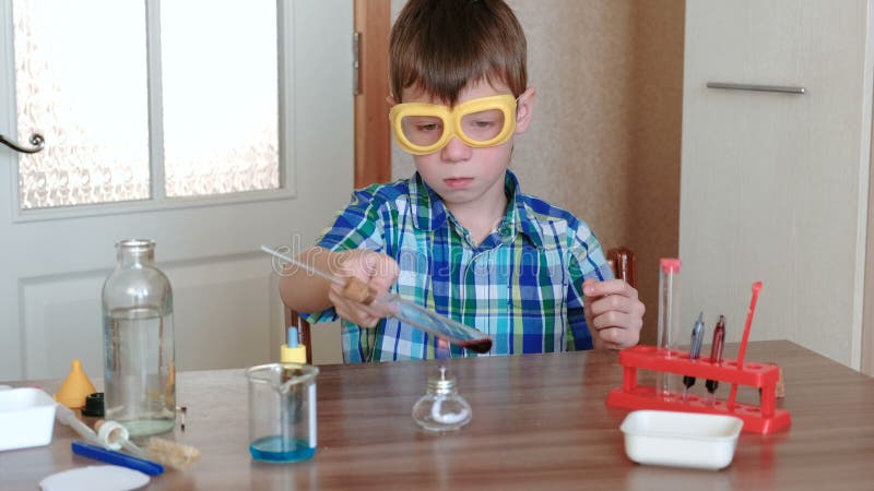 Experiências na química em casa O menino aquece o tubo de ensaio com líquido vermelho em lâmpada ardente do álcool