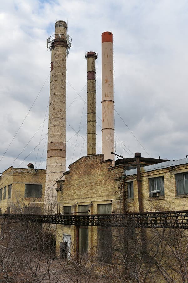 Old factory building with weathered yellow brick walls and large windows. Three tall chimneys, varying in height and color, rise prominently from the structure, exhibiting signs of rust. The sky is overcast, casting a grey tone over the scene. Metal scaffolding and pipes are visible, indicating industrial use. The foreground features sparse vegetation, and the overall environment appears neglected, suggesting abandonment or reduced activity. Sparse use stock images, royalty-free photos and pictures