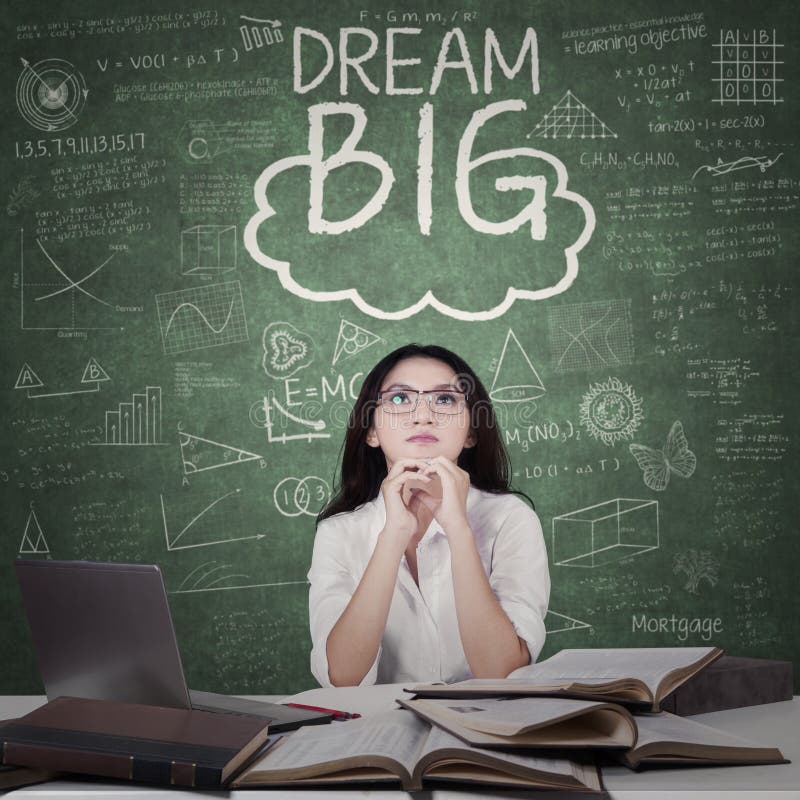 Pretty female high school student sitting in the classroom with books on the table and Dream Big text on the chalkboard. Pretty female high school student sitting in the classroom with books on the table and Dream Big text on the chalkboard