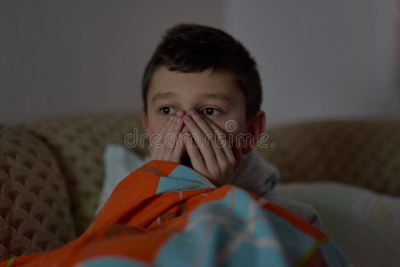 F?rskr?ckt barn som h?ller ?gonen p? l?skig film p? tv som sitter p? soffan p? natten royaltyfri bild F?rskr?ckt barn som h?ller ?gonen p? l?skig film p? tv som sitter p? soffan p? natten royaltyfri bild