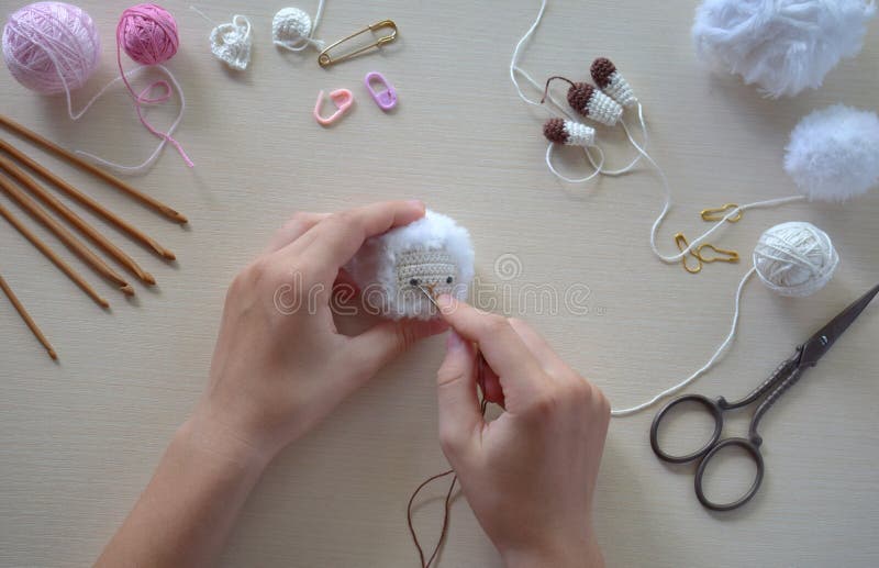 Making fanny sheep with wreath of flowers. Crochet toy ewe for child.  On table threads, needles, hook, cotton yarn. Step 2 - glue eyes of toy. Handmade crafts. DIY concept. Small business. Income from hobby. Making fanny sheep with wreath of flowers. Crochet toy ewe for child.  On table threads, needles, hook, cotton yarn. Step 2 - glue eyes of toy. Handmade crafts. DIY concept. Small business. Income from hobby