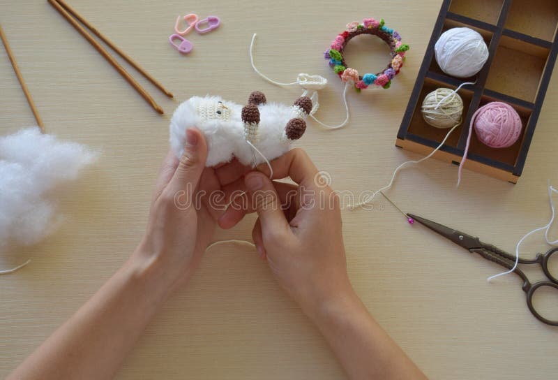 Making fanny sheep with wreath of flowers. Crochet toy ewe for child.  On table threads, needles, hook, cotton yarn. Step 2 - to sew all details of toy. Handmade crafts. DIY concept. Small business. Income from hobby. Making fanny sheep with wreath of flowers. Crochet toy ewe for child.  On table threads, needles, hook, cotton yarn. Step 2 - to sew all details of toy. Handmade crafts. DIY concept. Small business. Income from hobby