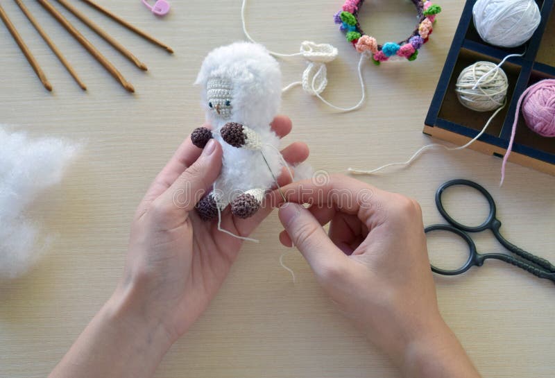 Making fanny sheep with wreath of flowers. Crochet toy ewe for child.  On table threads, needles, hook, cotton yarn. Step 2 - to sew all details of toy. Handmade crafts. DIY concept. Small business. Income from hobby. Making fanny sheep with wreath of flowers. Crochet toy ewe for child.  On table threads, needles, hook, cotton yarn. Step 2 - to sew all details of toy. Handmade crafts. DIY concept. Small business. Income from hobby