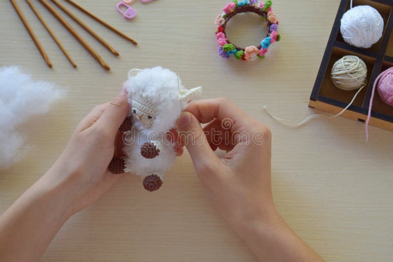 Making fanny sheep with wreath of flowers. Crochet toy ewe for child.  On table threads, needles, hook, cotton yarn. Step 2 - to sew all details of toy. Handmade crafts. DIY concept. Small business. Income from hobby. Making fanny sheep with wreath of flowers. Crochet toy ewe for child.  On table threads, needles, hook, cotton yarn. Step 2 - to sew all details of toy. Handmade crafts. DIY concept. Small business. Income from hobby