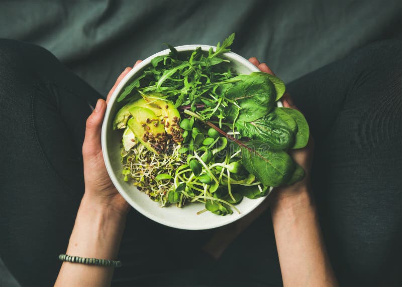 Green vegan breakfast meal in bowl with spinach, arugula, avocado, seeds and sprouts. Girl in leggins holding plate with hands visible, top view. Clean eating, dieting, vegan food concept. Green vegan breakfast meal in bowl with spinach, arugula, avocado, seeds and sprouts. Girl in leggins holding plate with hands visible, top view. Clean eating, dieting, vegan food concept